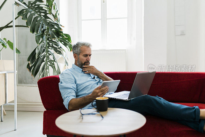 一个成熟的男人正坐在客厅的沙发上，他正在使用数码平板电脑和笔记本电脑