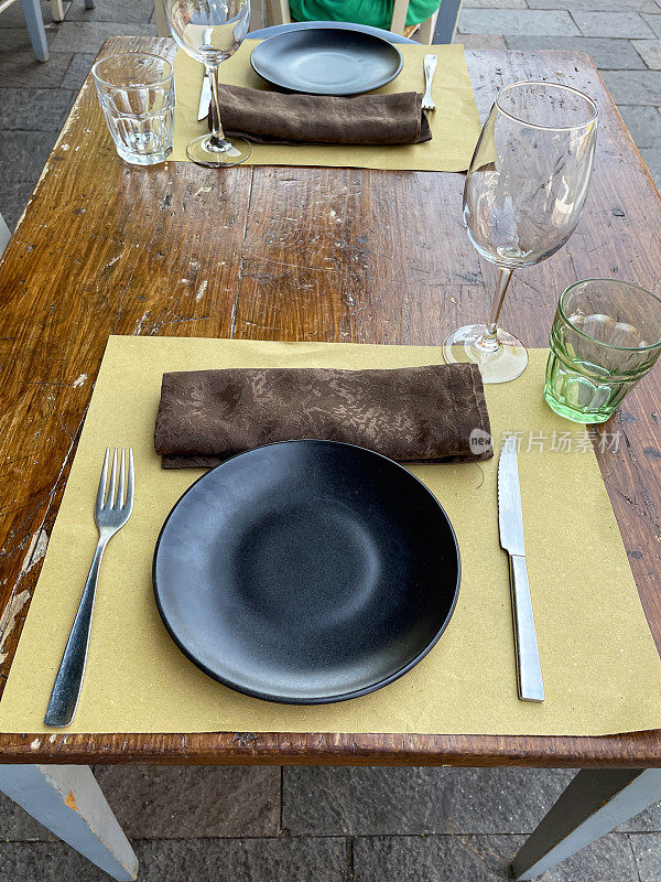 方形的特写图片，露天用餐区的木桌，两个地方的设置准备吃饭，空的，黑色的盘子放在桌垫上，棕色的，折叠的，材料餐巾，水杯，刀叉，酒杯，抬高的视图