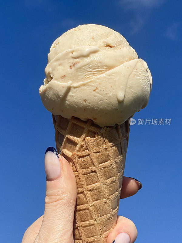 全画幅照片中，一个无法辨认的女人拿着华夫蛋筒，一勺太妃冰淇淋，黑色的指甲艺术，阳光明媚，晴朗的蓝天，聚焦在前景上
