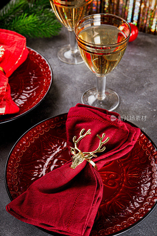 圣诞餐桌设置，餐巾上有金色的戒指鹿放在红色的盘子和两杯香槟在桌子上垂直的照片