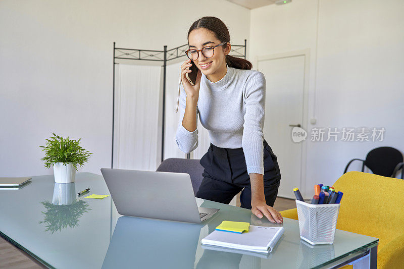 戴着眼镜的漂亮白人女孩在联合办公中心的私人办公室里工作，一边用电脑一边用智能手机和客户聊天