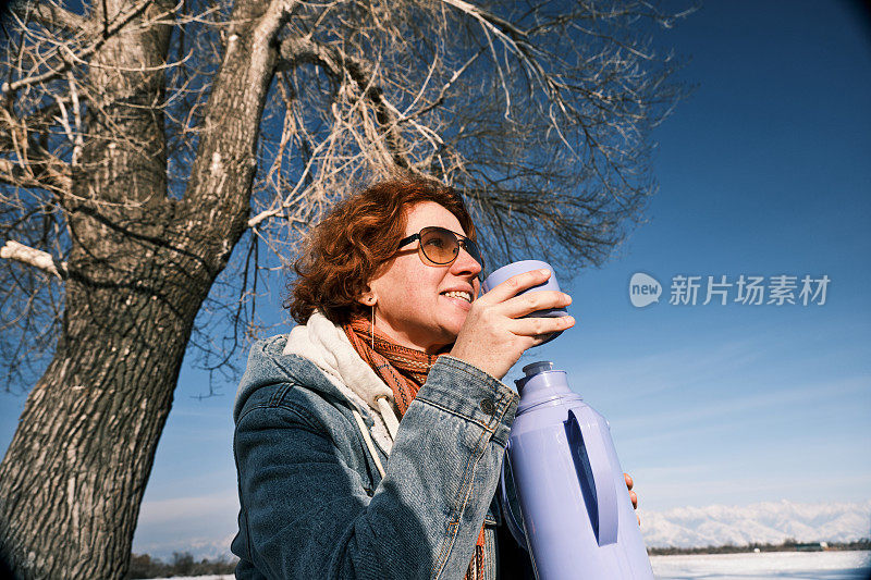 单身的成熟女游客坐在冬湾岸边的树桩上喝着热茶