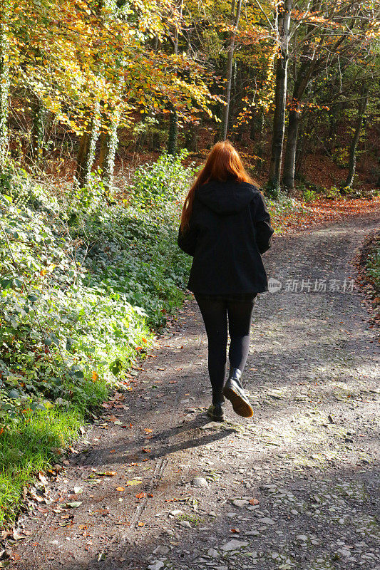 这是一个难以辨认的红发少女在秋天的阳光下走在英国乡村林地的荫凉小路上，昏暗的柏油马路，秋天彩色的树叶，光秃秃的树枝，冠状病毒封锁演练