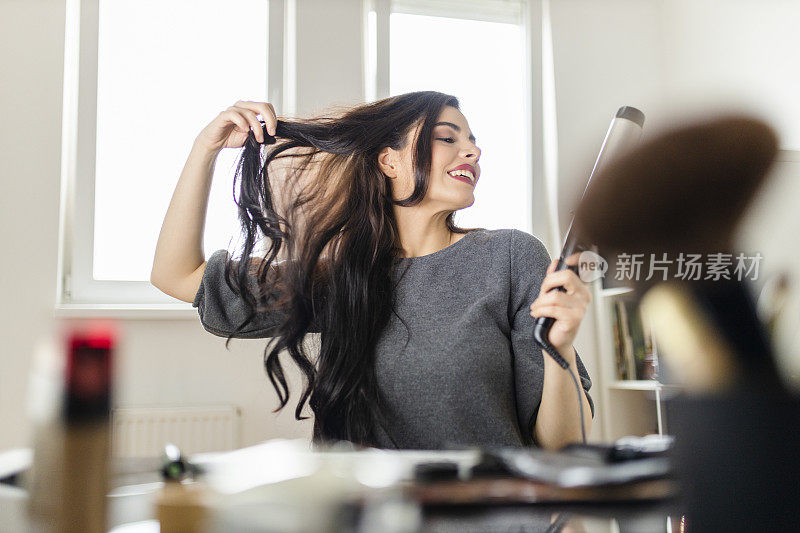 坐在梳妆台边看电脑边用卷发棒的女人。在冠状病毒大流行的时候，一名妇女通过在网上看教程来做头发