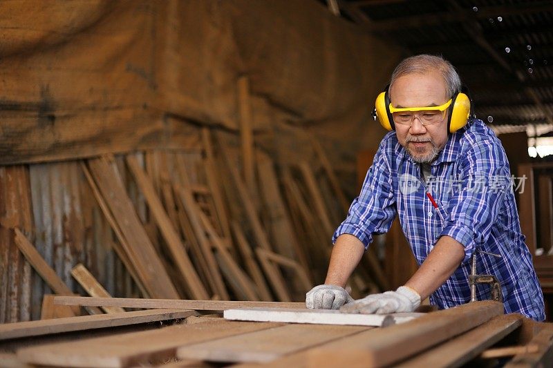年长的亚洲木匠正在他自己的车库风格的车间里锯木板在退休年龄的爱好