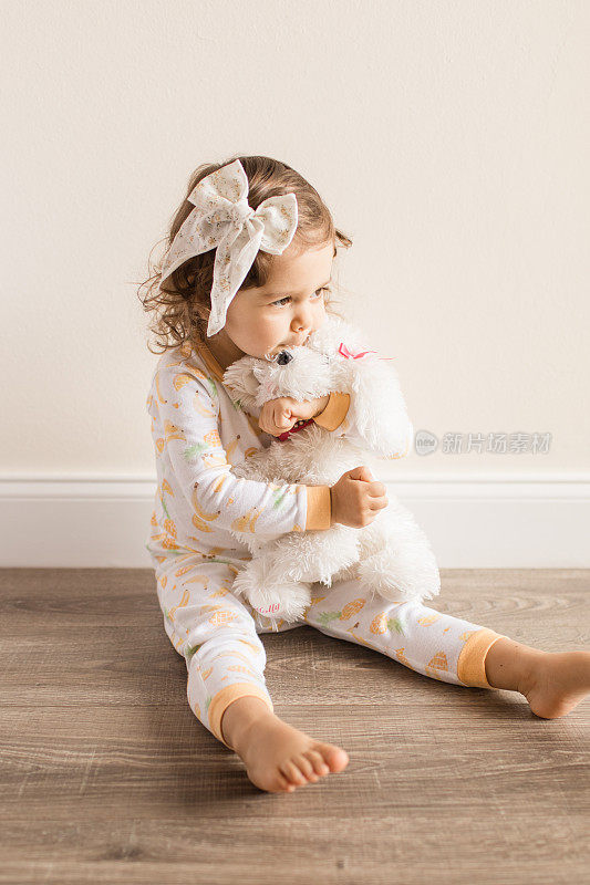 2019冠状病毒病期间，两岁蹒跚学步的小女孩穿着睡衣和蝴蝶结在家拥抱和亲吻她毛茸茸的白色狗玩具
