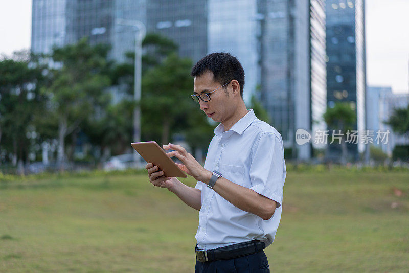 一位亚洲男性白领正在办公大楼外的草地上使用平板电脑