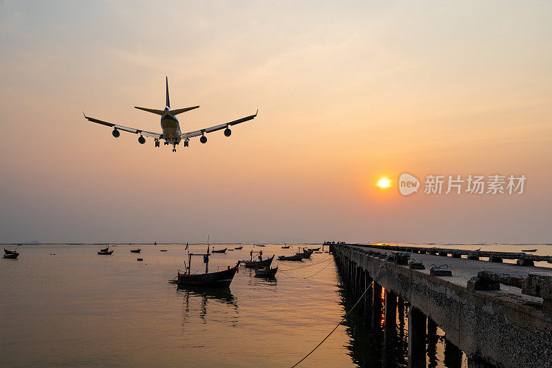 后方影像商业客机或货机飞过渔舟，在码头的海面上漂浮着金色的日落海景