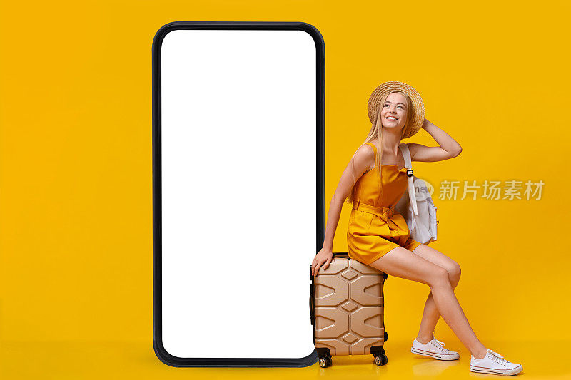 可爱的金发女郎坐在行李箱上，旁边是一个巨大的智能手机模型，正在网上预订暑假