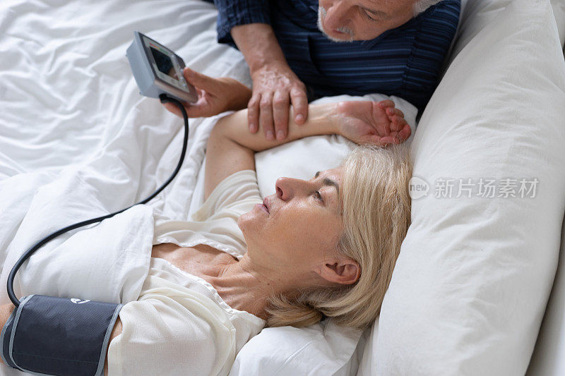 一对老夫妇早上躺在床上，妻子感觉不舒服，丈夫在测量她的血压