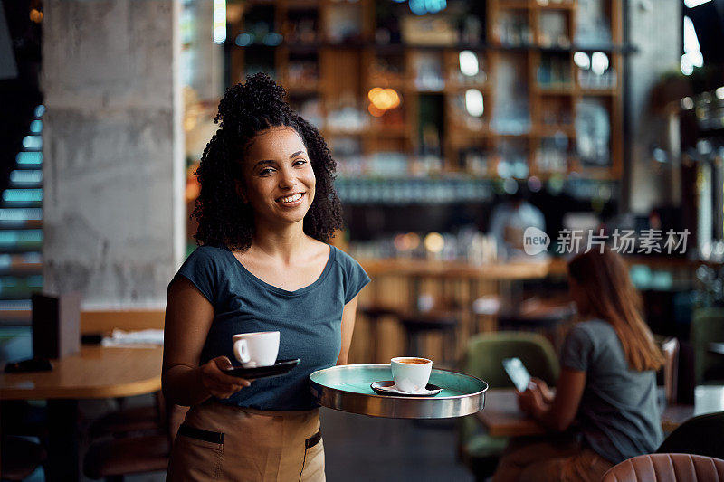 快乐的黑人女服务员在咖啡馆提供咖啡，看着相机。