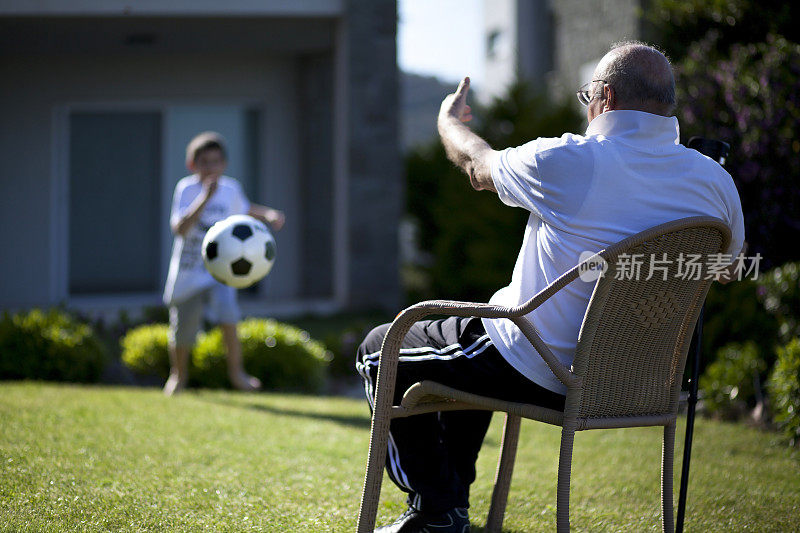爷爷坐在椅子上，他的孙子在踢足球