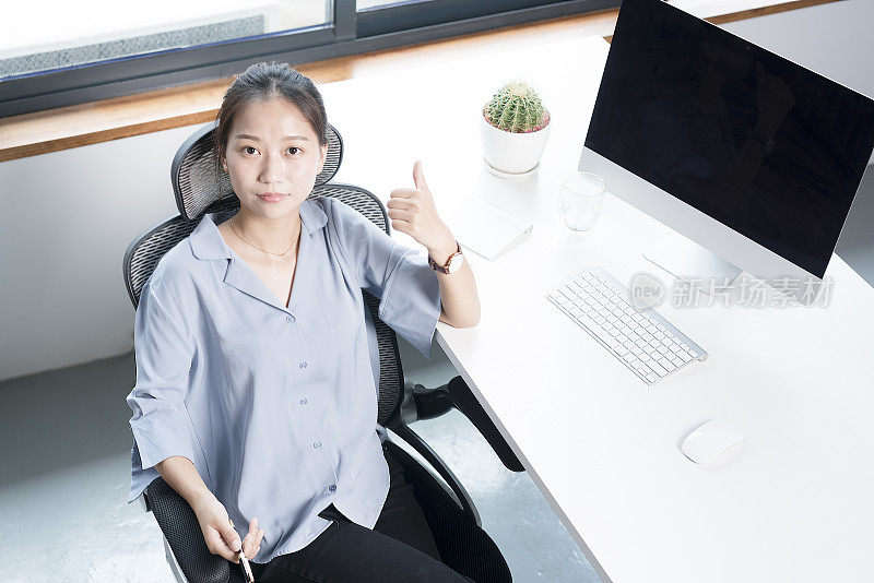 年轻女子坐在办公桌旁，对着镜头竖起大拇指