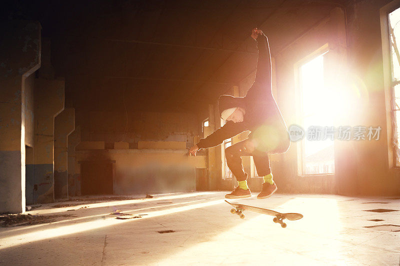 一个年轻的溜冰者戴着白色的帽子，穿着黑色的运动衫，在夕阳的背光下，在一座废弃的建筑里表演了一个溜冰跳的把戏
