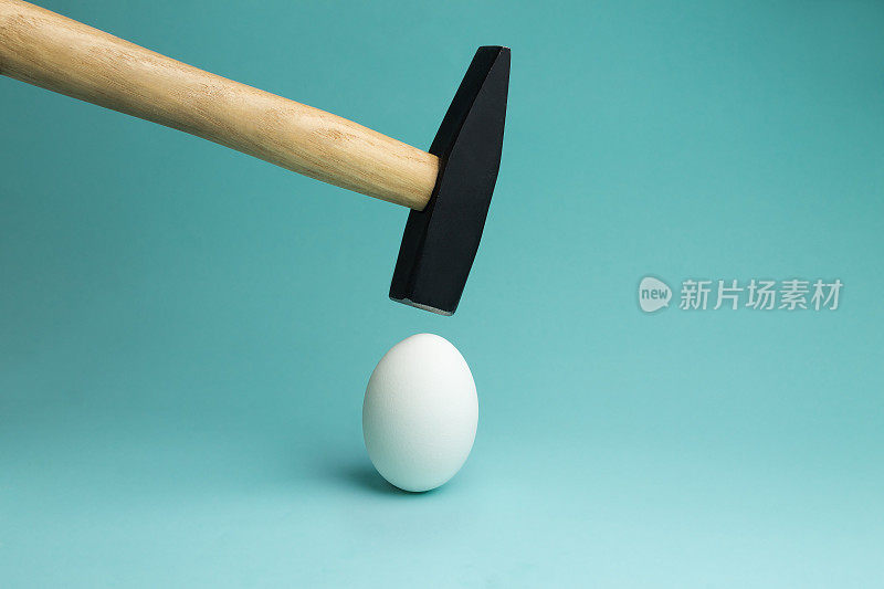 鸡蛋和锤子在它上面盘旋，在撞击之前。一个鸡蛋，用锤子在蓝色背景上敲