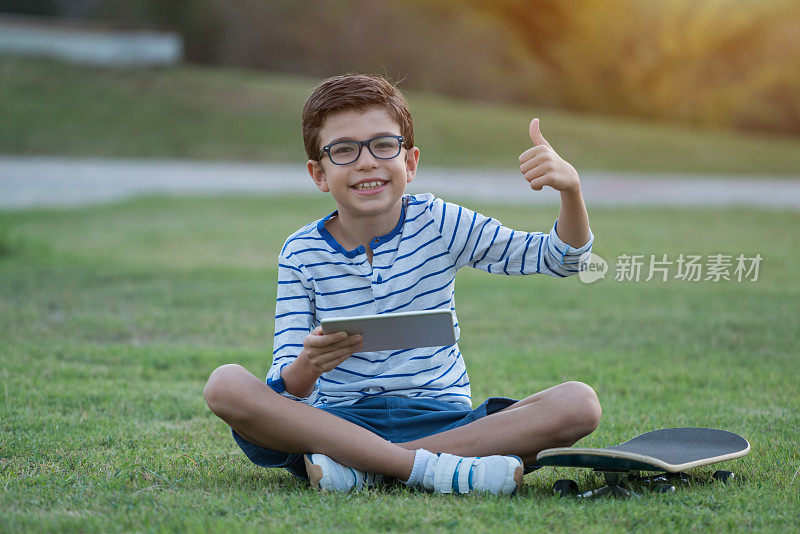 拿着平板电脑和滑板的男孩坐在草地上