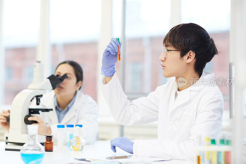 年轻认真的亚洲实验室员工穿着白大褂坐在桌子旁，拿着实验室工具，看着试管里的液体