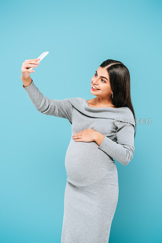 微笑的怀孕漂亮女孩在智能手机上自拍孤立在蓝色