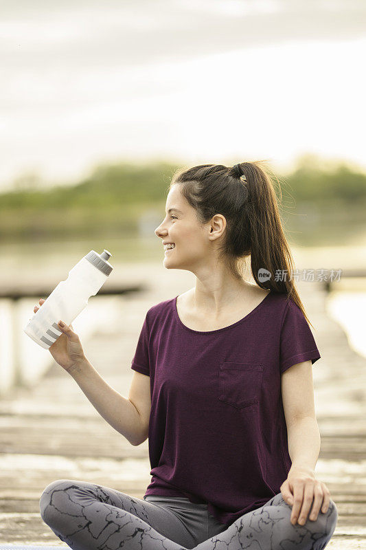 喝着水坐在湖边码头上的运动型女人