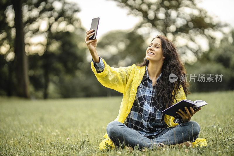 穿着黄色雨衣的快乐女人，用手机自拍，在大自然中读书