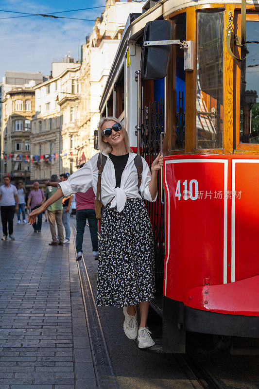 土耳其伊斯坦布尔，Istiklal大街上，一辆怀旧的红色Istiklal电车的运行板上挂着一个背包，穿着白衬衫和裙子的年轻金发美女。垂直图