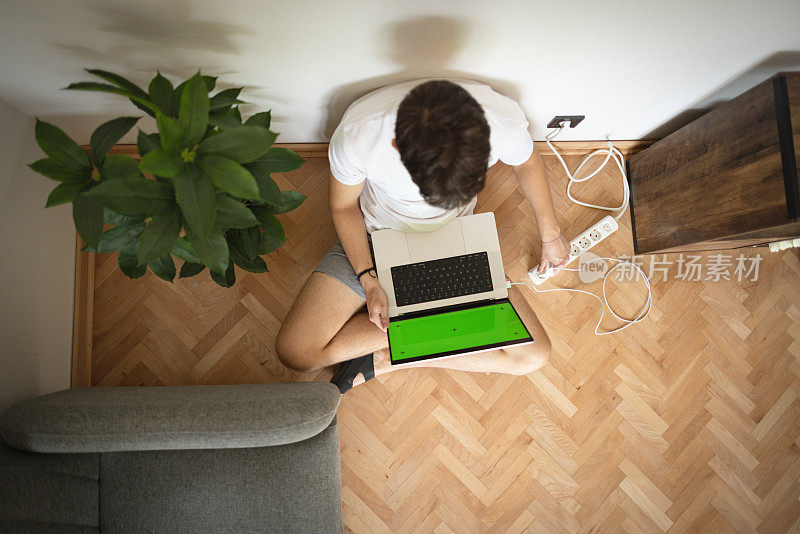 一个十几岁的男孩，坐在地板上，把笔记本电脑充电器插到延长线上