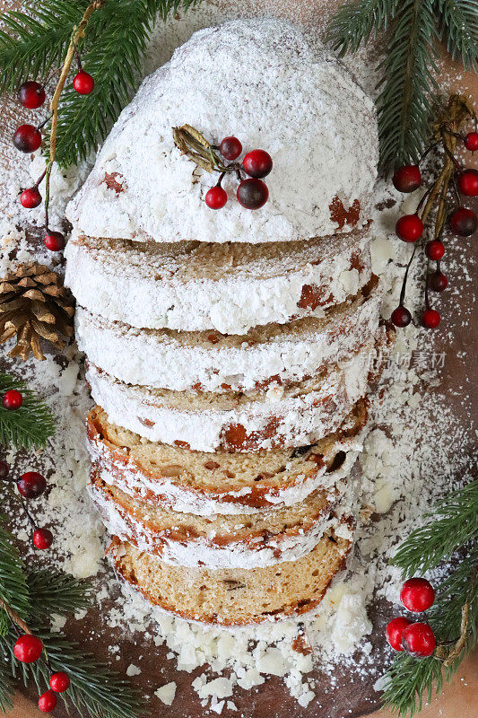 切片的圣诞蛋糕，甜面包片，杏仁糖，干果，包括用糖粉装饰的醋栗和葡萄干，松果，云杉针，红色浆果，棕色背景，高架视图