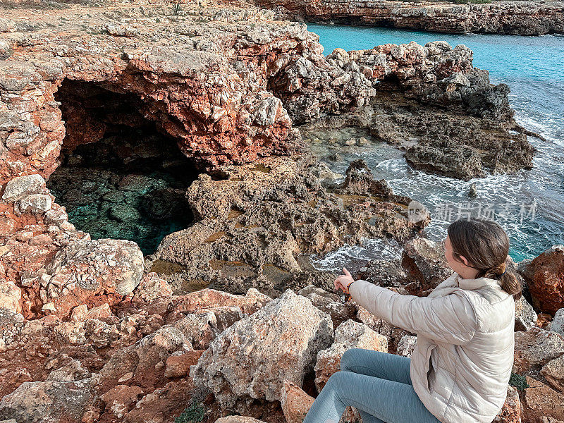 一个十几岁的女孩坐在悬崖的岩石上，指着一个海洞