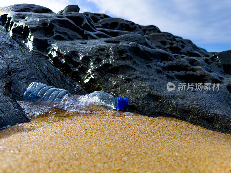 在葡萄牙的海滩上，一个装满垃圾的空塑料瓶被粗心地遗忘在一块岩石旁边的沙滩上