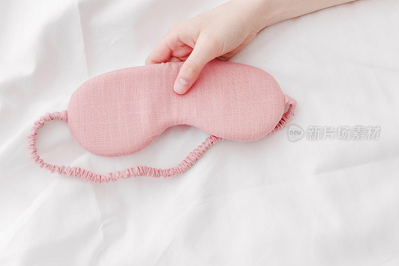 粉红色的眼罩睡在女人的手在白色的床上，最小的生活方式美学的照片。俯视图女性睡眠面膜，适合最佳睡眠者，适合旅行，舒适放松。好好休息的概念。