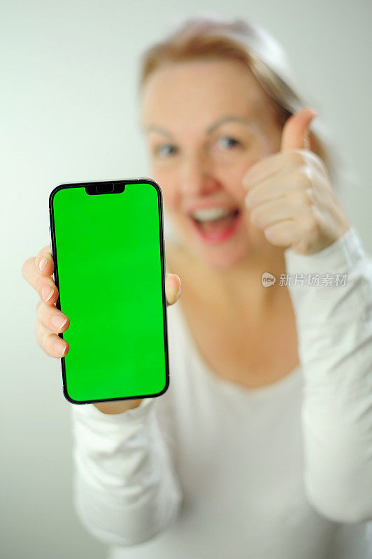 竖起大拇指展示类chromakey绿屏广告照片兴奋兴奋的女士手持新款智能手机呈现优质触摸屏设备张开嘴穿白色夹克