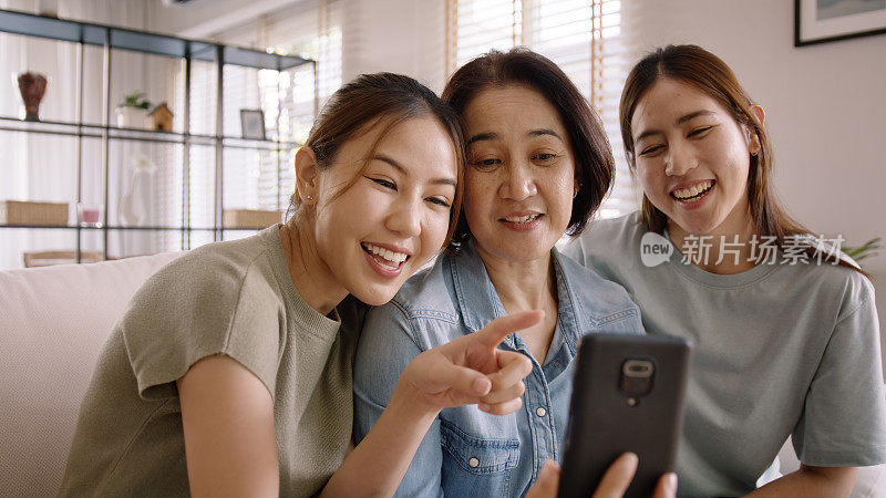 一群亚洲人，年轻的成年妇女和中年妈妈看着网络摄像头，在网上视频聊天，舒适地坐在家里的沙发上。妈妈和孩子一起享受放松的微笑自拍。