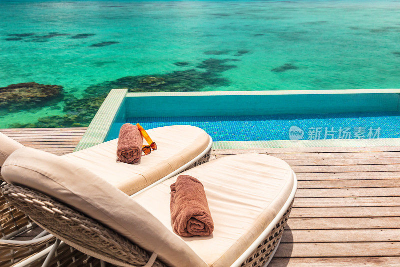 豪华酒店无限游泳池，甲板上有两个日光浴躺椅，配有毛巾，情侣蜜月放松水上平房套房。海洋暗礁蓝色背景。