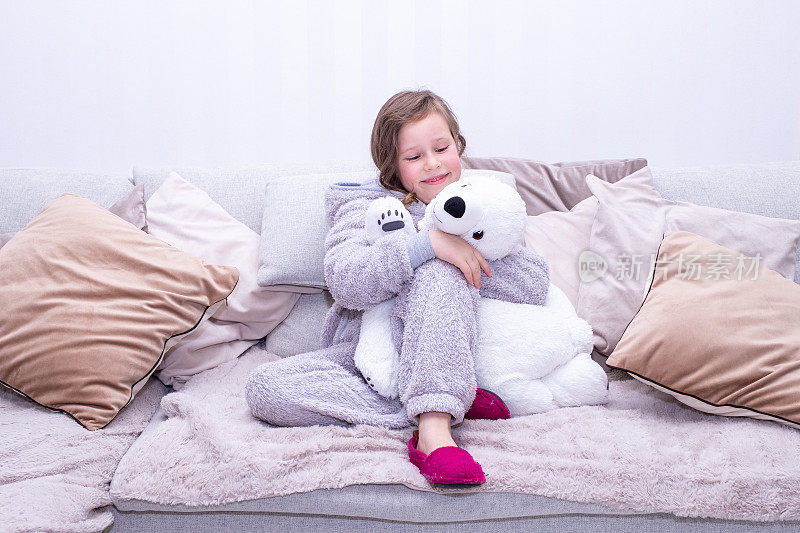 一幅白人儿童的肖像，一个7岁的女孩穿着长毛绒工作服，坐在柔软的大沙发上，抱着一个柔软的玩具——北极熊。