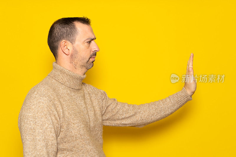 拒绝，否认的概念。穿着高领毛衣的男子侧视图伸出手做停止标志，用否定的手势发出警告。室内工作室拍摄孤立的黄色背景