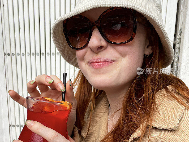 有魅力的红发女子从沾满塑料吸管、冰块和橘子片的玻璃杯中啜着鸡尾酒，在户外戴着灯芯绒遮阳帽和夹克，戴着太阳镜，重点放在前景上