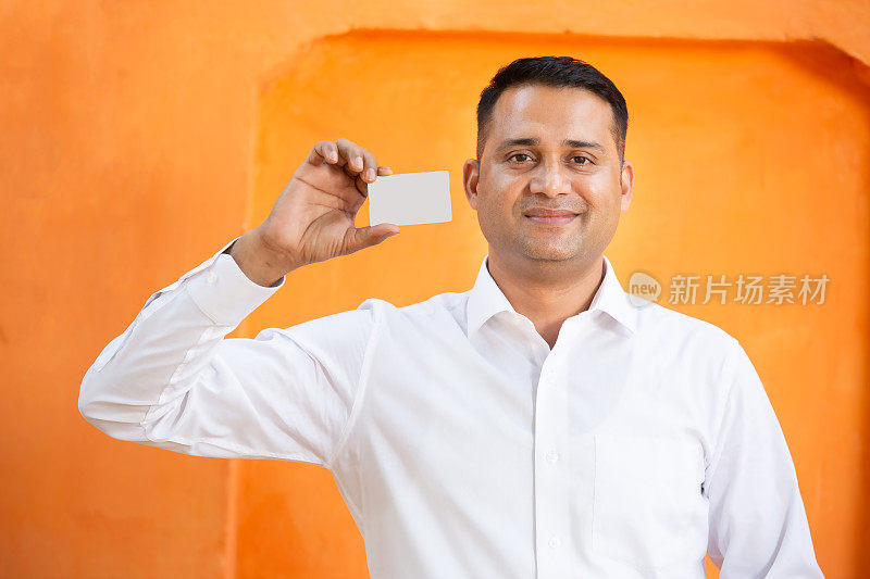 年轻的印度男子在橙色背景下展示空白的白色卡片模型，微笑的亚洲男子拿着空白的商务卡或电子卡，用于支付、借记卡、信用卡、银行，选择性聚焦。