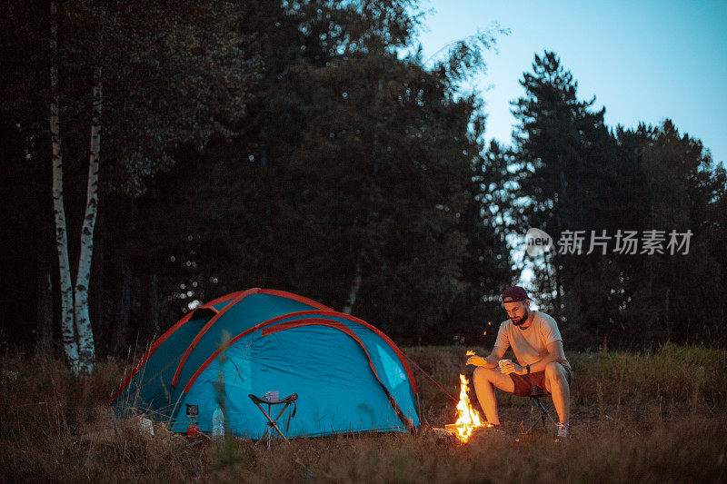 年轻的徒步旅行者坐在露营椅旁的火和露营帐篷和喝咖啡的塑料杯在大自然中