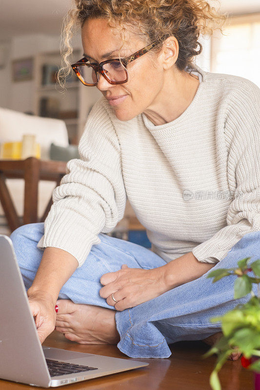 一位中年女士坐在地板上舒舒服服地使用笔记本电脑独自在室内科技无线休闲活动。现代女性在电脑上工作。女人浏览网页和写作