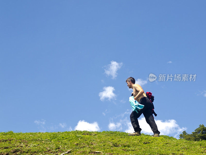 赤裸上身的男性徒步者在阳光下爬一个长满草的小山。