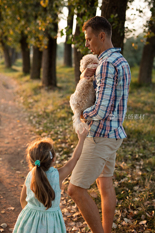 可爱的孩子问她爸爸让狗趴在地上，这样她就可以和他玩了。爸爸，女儿和狗在公园里散步。