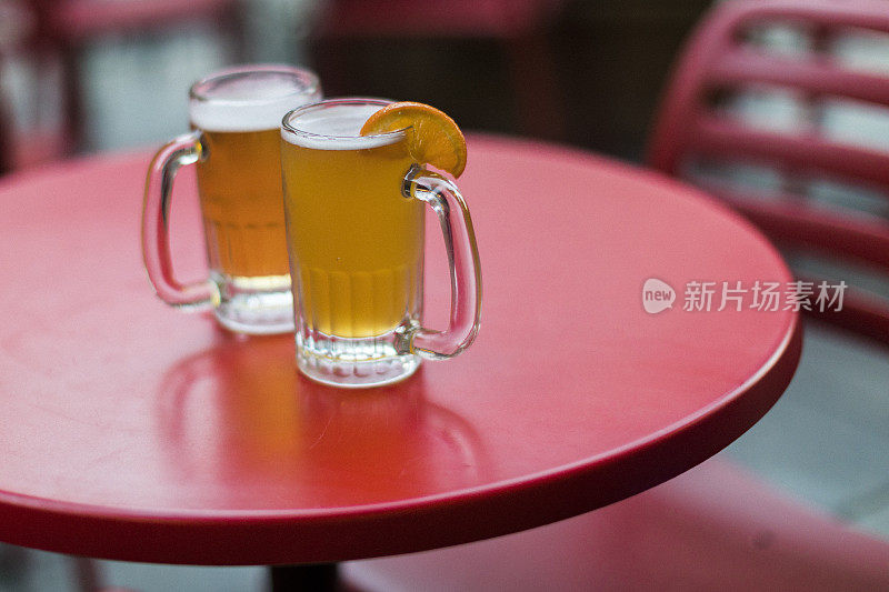 两个清爽的啤酒杯放在酒吧露台的红桌上，一个是杏子啤酒，另一个是奶油啤酒。