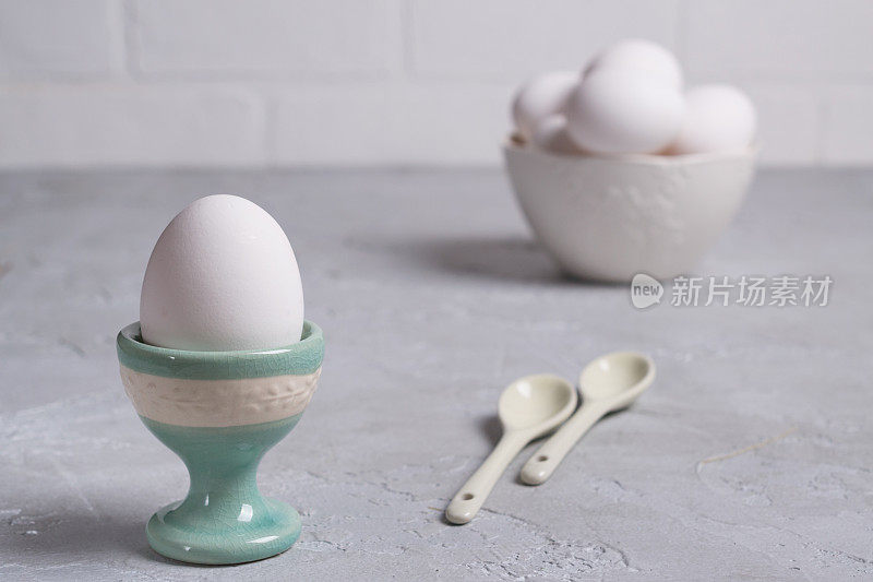 白色的鸡蛋在陶瓷支架和鸡蛋在白色的碗。在灰色的混凝土背景上。