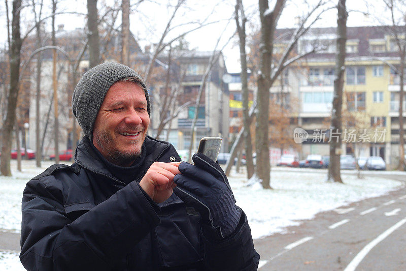 英俊快乐的成年人在下雪的冬天在城市街道上使用手机