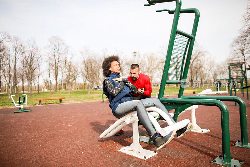 年轻的女运动员在公园的长椅上做仰卧起坐，健身教练在测量她的时间并监视她
