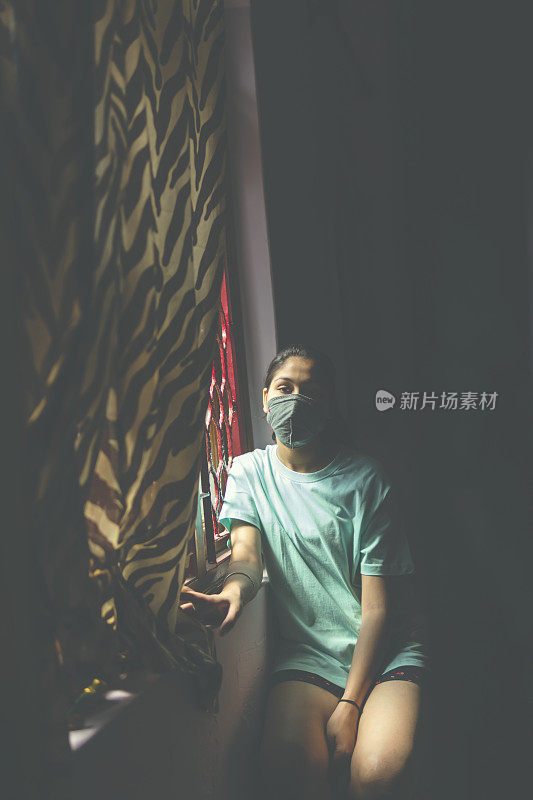 这个悲伤的女孩正在保护自己，戴着口罩，在家里自我隔离。