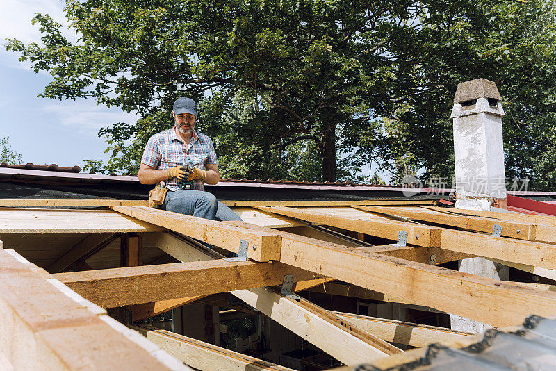 阳光明媚的日子里，成熟的男性木匠拿着电动工具坐在木屋顶上靠树