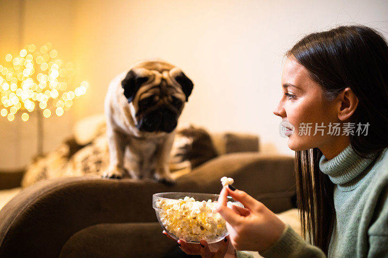 一个年轻漂亮的女人在吃爆米花，一只可爱的哈巴狗在盯着她看