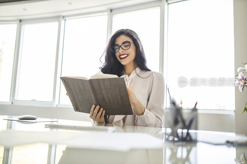 年轻的商业女性在她的办公室里阅读日记，摇摇一天，在电脑上工作