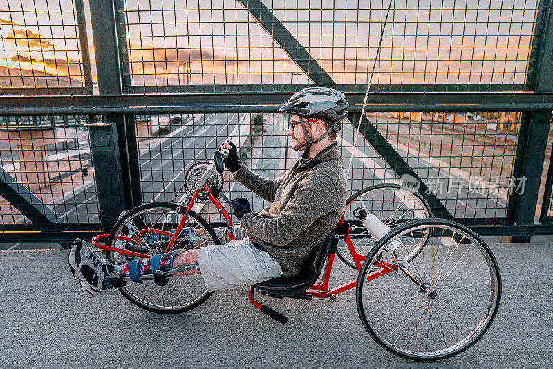 一个帅气的年轻人在日落时分骑着一辆红色的自行车，穿着矫形的腿部支架，在城市的人行天桥上进行户外适应性锻炼
